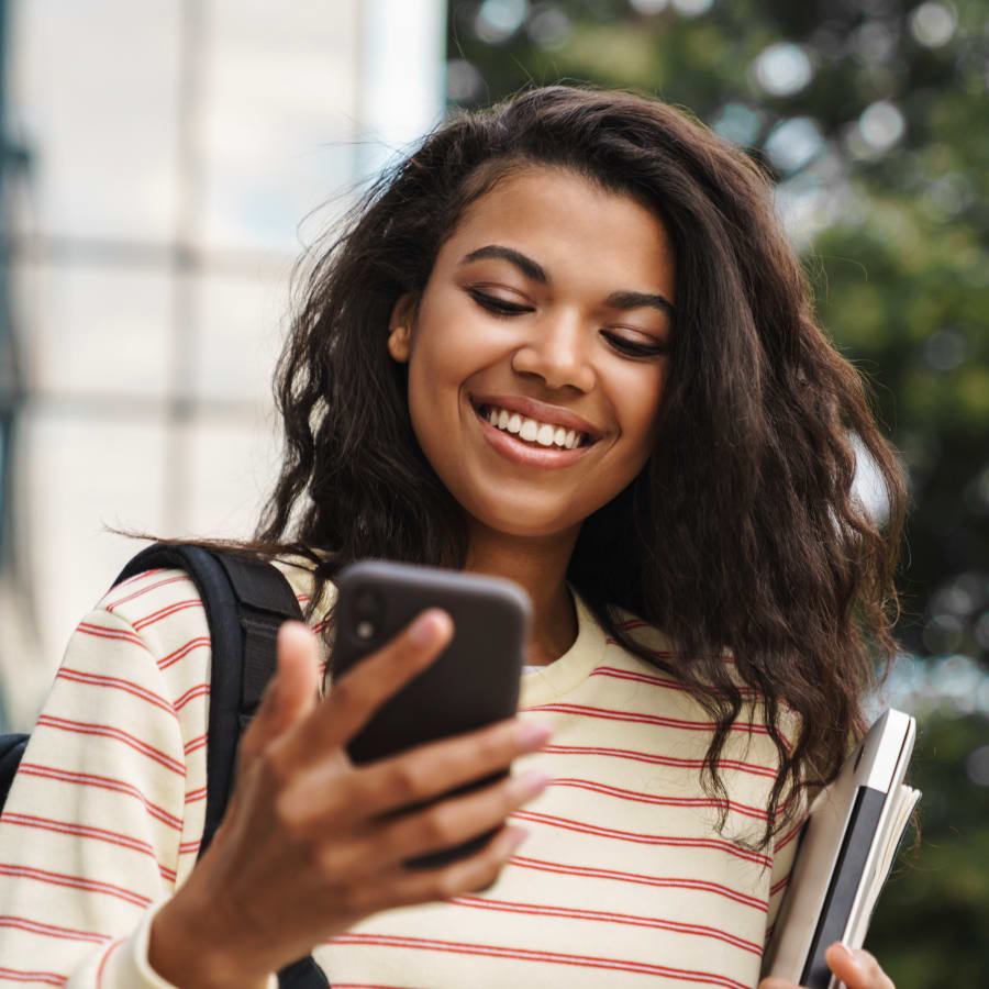 A teenage girl smiles at her phone. She is standing outside holding a laptop and some books.